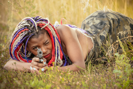 portrait of a beautiful young African American girl with a dreddy hairdress on a park background, nature, deciduous forest, with a gun in her hands. concept of photo walks, female self-defenseの写真素材