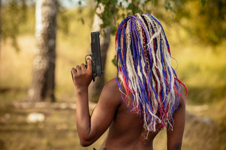 portrait of a beautiful young African American girl with a dreddy hairdress on a park background, nature, deciduous forest, with a gun in her hands. concept of photo walks, female self-defenseの写真素材
