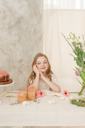 A girl with long hair in a light dress is sitting at the Easter table with cakes, spring flowers and...
