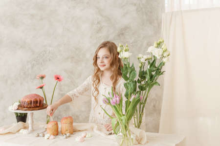A girl with long hair in a light dress is sitting at the Easter table with cakes, spring flowers and quail eggs. Happy Easter celebration.の写真素材