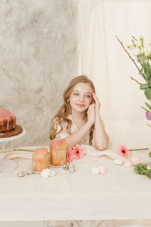 A girl with long hair in a light dress is sitting at the Easter table with cakes, spring flowers and quail eggs. Happy Easter celebration.の写真素材