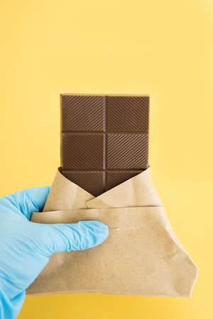 Bar of milk chocolate in a wrapper is held by a hand in a medical glove on a yellow background. Sweetness. World chocolate day 2020 during the coronavirus.の写真素材