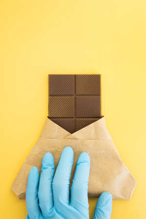 Bar of milk chocolate in a wrapper is held by a hand in a medical glove on a yellow background. Sweetness. World chocolate day 2020 during the coronavirus.の写真素材