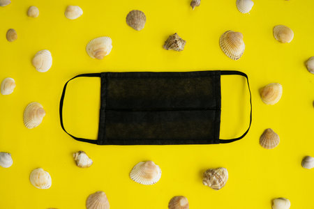 Black medical face mask and seashells on a yellow background. Flat lying, copy space, holiday...