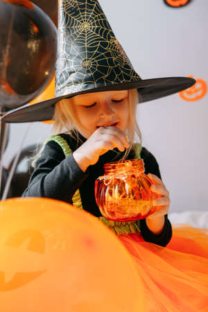 Childrens Halloween - a girl in a witch hat and a carnival costume with airy orange and black balloons at home. Ready to celebrate Halloweenの写真素材