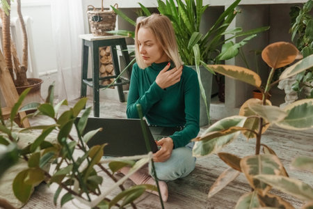 A blonde woman in a room with a lot of green indoor plants is working on a laptop. The concept of...