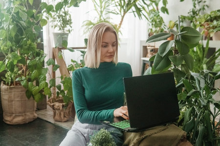 A blonde woman in a room with a lot of green indoor plants is working on a laptop. The concept of...