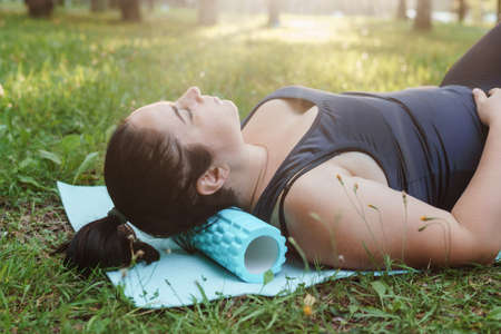 A charming brunette woman plus-size body positive practices sports in nature. Woman is engaged with a massage roller for the body in the park on a sports mat.の写真素材