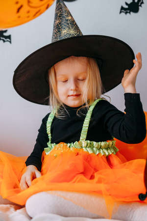 Childrens Halloween - a girl in a witch hat and a carnival costume with airy orange and black balloons at home. Ready to celebrate Halloweenの写真素材