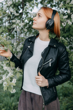 A red-haired teenage girl listens to music in headphones, configured via the phone. The time of spring flowering of cherry orchardsの写真素材