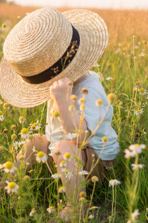 A little blond boy in a straw hat is sitting in the grass in a chamomile field. The concept of walking in nature, freedom and an environmentally friendly lifestyle.の写真素材