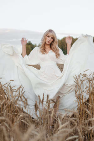 A blonde woman in a long white dress walks in a wheat field. The concept of a wedding and walking in natureの写真素材