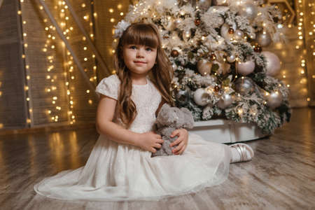A girl in a festive light dress with a plush toy in her hands next to a Christmas tree, lights of garlands in the background. The concept of New Year holidaysの写真素材