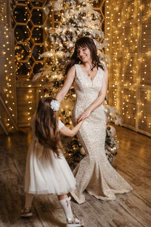 A little girl with her mother in light festive dresses next to the Christmas tree. The theme of New Years holidays and festive interior with garlands and light bulbsの写真素材