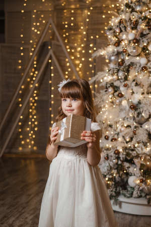 A girl in a festive light dress with a gift in her hands next to a Christmas tree, lights of garlands in the background. The concept of New Year holidaysの写真素材