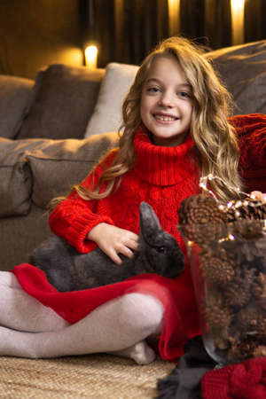 A little blonde girl with a gray rabbit in her arms next to fireplace. The concept of winter holidayの写真素材