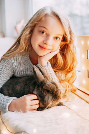 A beautiful blonde girl is sitting on the windowsill with a gray rabbit in a Christmas atmosphere.の写真素材