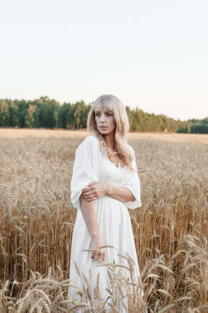 A blonde woman in a long white dress walks in a wheat field. The concept of a wedding and walking in natureの写真素材