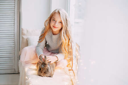 A beautiful blonde girl is sitting on the windowsill with a gray rabbit in a Christmas atmosphere.の写真素材