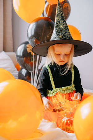 Childrens Halloween - a girl in a witch hat and a carnival costume with airy orange and black balloons at home. Ready to celebrate Halloweenの写真素材