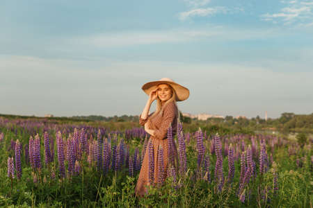 A beautiful woman in a straw hat walks in a field with purple flowers. A walk in nature in the lupin fieldの写真素材
