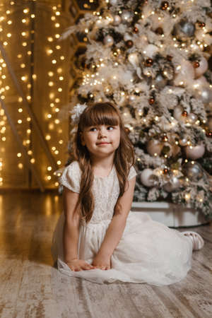 A girl in a festive light dress next to a Christmas tree, lights of garlands in the background. The concept of New Year holidays.の写真素材