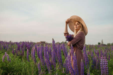 A beautiful woman in a straw hat walks in a field with purple flowers. A walk in nature in the lupin fieldの写真素材