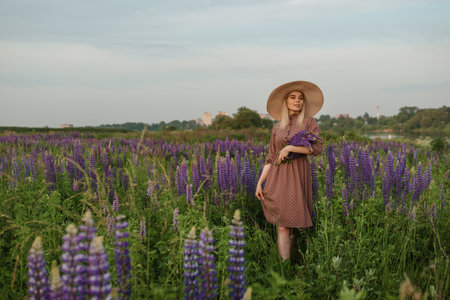 A beautiful woman in a straw hat walks in a field with purple flowers. A walk in nature in the lupin fieldの写真素材