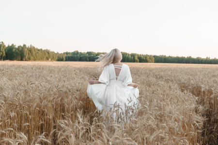 A blonde woman in a long white dress walks in a wheat field. The concept of a wedding and walking in natureの写真素材