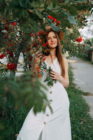 A beautiful blonde girl in a hat with long hair, on a walk in the autumn season. Portrait of a woman at a rowan tree.の写真素材