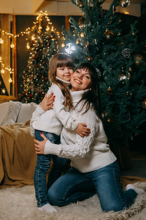 A little girl with her mother in a cozy home environment on the sofa next to the Christmas tree. The theme of New Year holidays and festive interior with garlands and light bulbsの写真素材