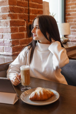 The brunette is working with a tablet at a table in a cafe. Drinks latte coffee and eats a croissant. The topic of freelancing and free work scheduleの写真素材