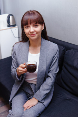 A brunette in a gray suit is sitting on the couch in the office with a cup of black coffee. Business conceptの写真素材