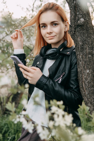 A red-haired teenage girl listens to music in headphones, configured via the phone. The time of spring flowering of cherry orchardsの写真素材