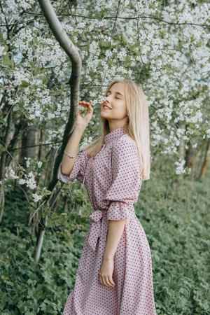 Blonde girl on a spring walk in the garden with cherry blossoms. Female portrait, close-up. A girl in a pink polka dot dress.の写真素材