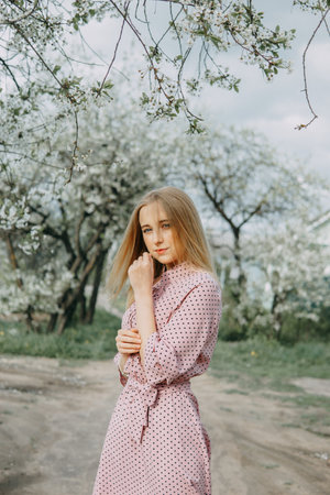 Blonde girl on a spring walk in the garden with cherry blossoms. Female portrait, close-up. A girl in a pink polka dot dress.の写真素材