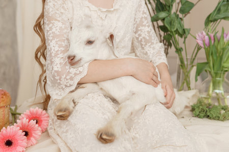 The girl is sitting at the Easter table with cakes, spring flowers and quail eggs and holding a white kid in her arms. Happy Easter celebrationの写真素材
