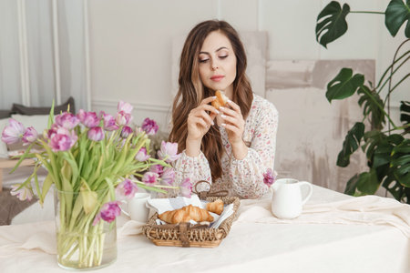 A brown-haired woman with long hair is having breakfast at the table with croissants and a cup of...