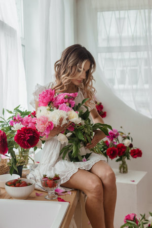 A beautiful girl in a white nightgown at home surrounded by spring flowers. A room decorated with bouquets of peonies.の写真素材
