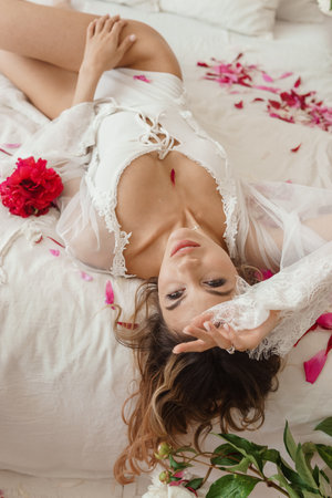 A beautiful girl in a white nightgown at home, surrounded by spring flowers on a large bed. A room decorated with bouquets of peoniesの写真素材