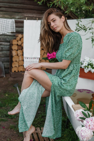 A woman is sitting on a cast-iron bathtub in the courtyard of a country house next to a bush of flowering peonies. The concept of summer, country life, a bathroom on the street in a blooming garden in the countryの写真素材