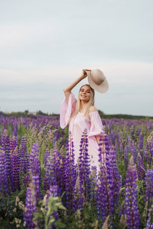 A beautiful woman in a straw hat walks in a field with purple flowers. A walk in nature in the lupin fieldの写真素材