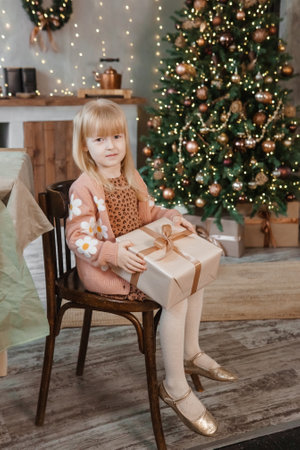 A little blonde girl is sitting on a wooden chair in a Scandinavian interior decorated in a New Years style. A child holds a Christmas gift in a craft package.の写真素材