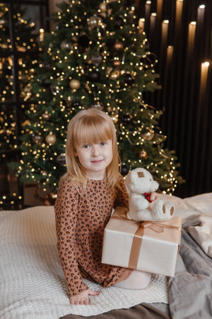 A little blonde girl is sitting on a soft bed in a festive interior decorated in a New Years style. A child holds a Christmas gift in a craft package.の写真素材
