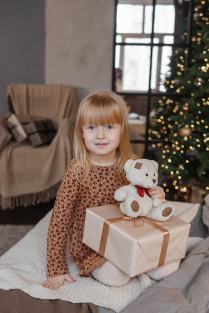 A little blonde girl is sitting on a soft bed in a festive interior decorated in a New Years style. A child holds a Christmas gift in a craft package.の写真素材