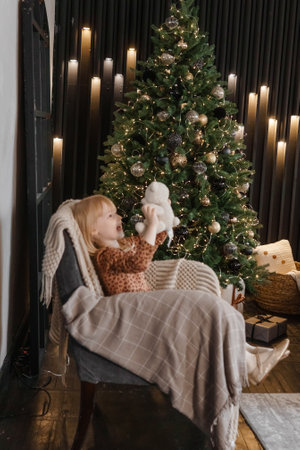 A little blonde girl is sitting in an armchair in an interior decorated in a New Years style. A child holds a teddy bear in his hands.の写真素材