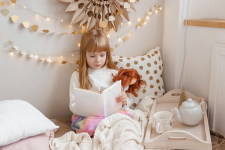 A little red-haired girl is reading a book to her doll in a nursery decorated for the Christmas holidays.の写真素材