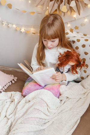 A little red-haired girl is reading a book to her doll in a nursery decorated for the Christmas holidays.の写真素材