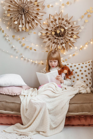 A little red-haired girl is reading a book to her doll in a nursery decorated for the Christmas holidays.の写真素材