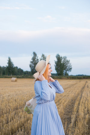 A red-haired woman in a hat and a blue dress walks in a field with haystacks.の写真素材